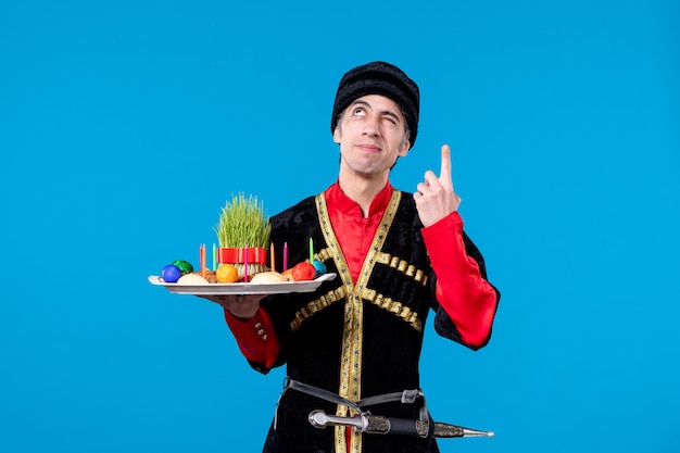 Front view of young thoughtful man in traditional dress holding tray filled with national confectionery on blue background