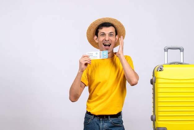Вид спереди молодой турист, стоящий возле желтого чемодана, выражая свои чувства с билетом