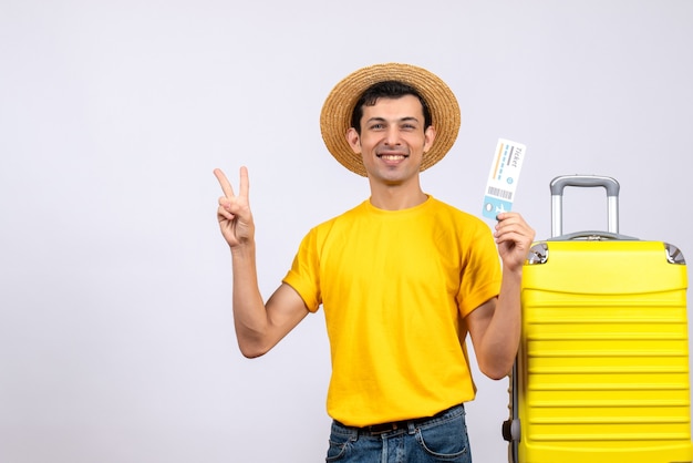 航空券を保持しているvサインを作る黄色のスーツケースの近くに立っている正面図若い観光客