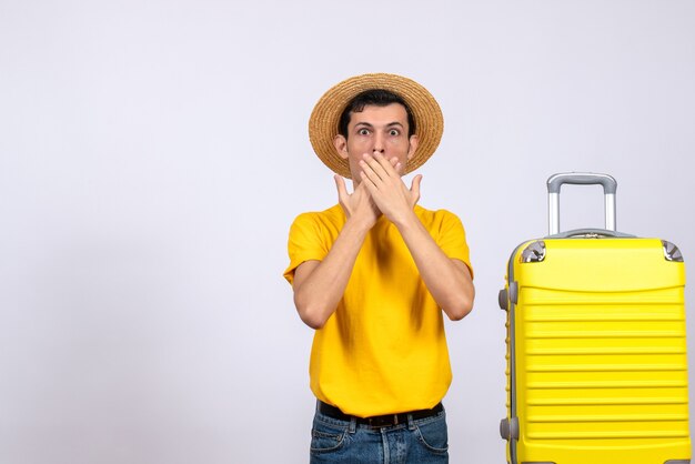 Вид спереди молодой турист, стоящий возле желтого чемодана, кладя руки ему в рот