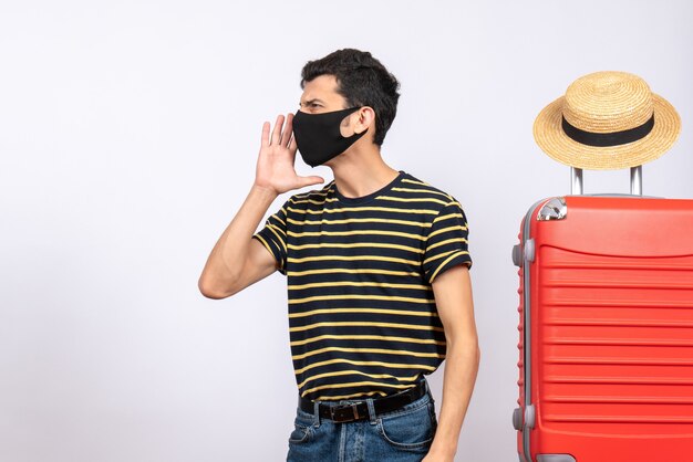 Вид спереди молодой турист с черной маской, стоящий возле красного чемодана, звонящий кому-то