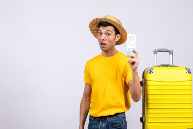 Вид спереди молодой турист в соломенной шляпе, стоящий возле желтого чемодана с билетом