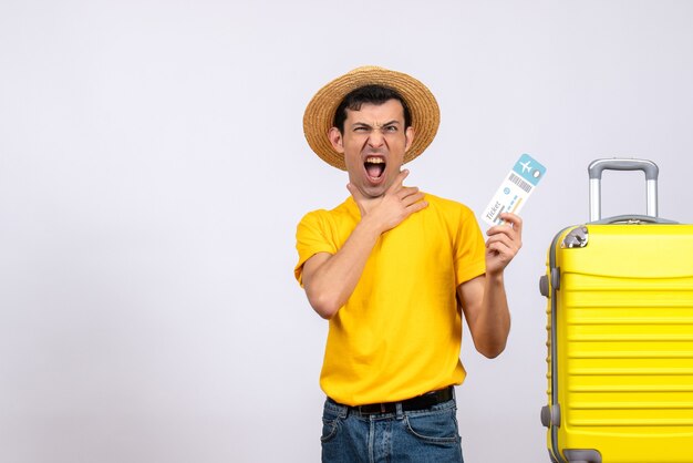 Front view young tourist in yellow t-shirt standing near yellow suitcase holding air ticket