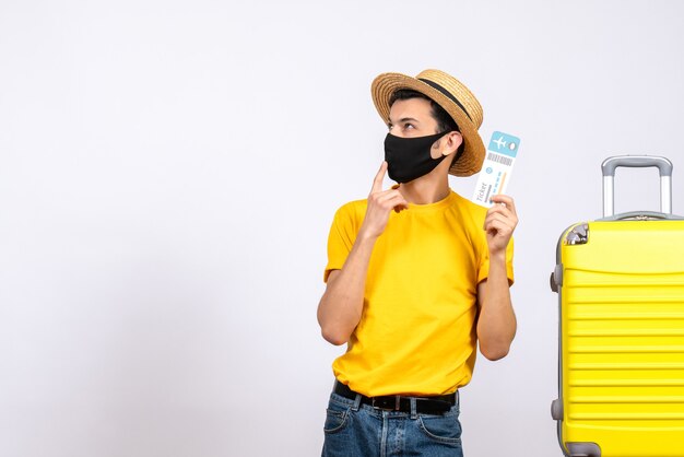 Front view young tourist in yellow t-shirt standing near yellow suitcase holding up air ticket