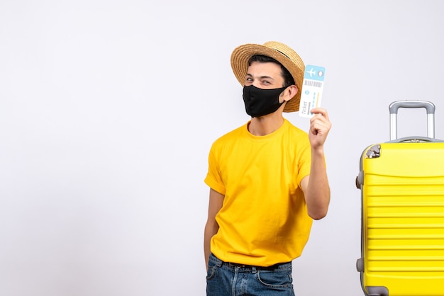 Front view young tourist in yellow t-shirt standing near yellow suitcase holding up travel ticket