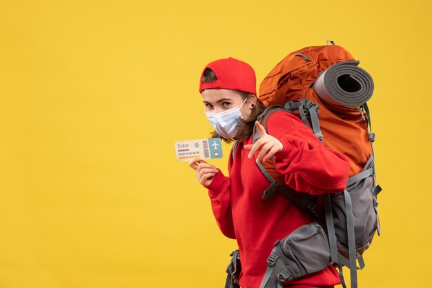 Front view young traveler girl with backpack and mask holding plane ticket pointing at camera