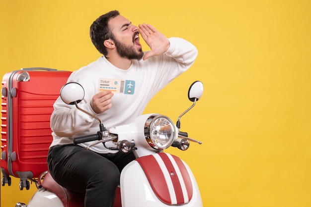 Front view of young travelling man sitting on motorcycle with suitcase on it holding ticket calling someone on isolated yellow background