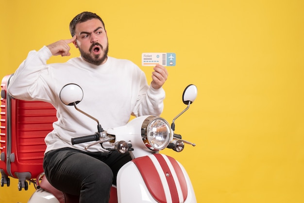 Front view of young uncertain unsure travelling man sitting on motorcycle with suitcase on it holding ticket on isolated yellow background