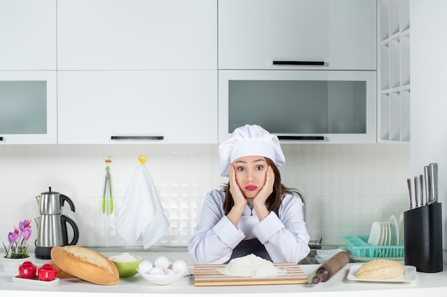 白いキッチンでまな板の食べ物とテーブルの後ろに立っている制服を着た若い不満と不確かな混乱した女性シェフの正面図