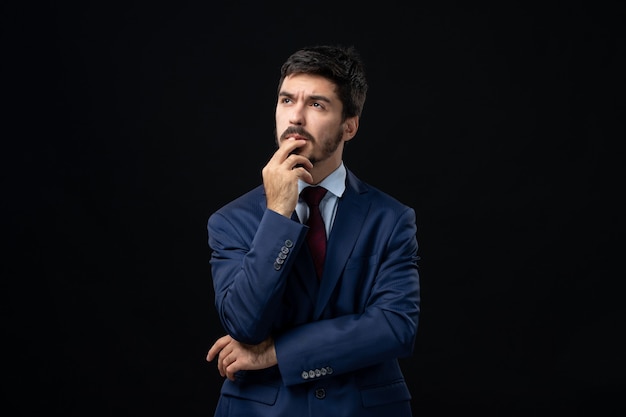 Front view of young and unsure bearded man in deep thoughts on isolated dark wall