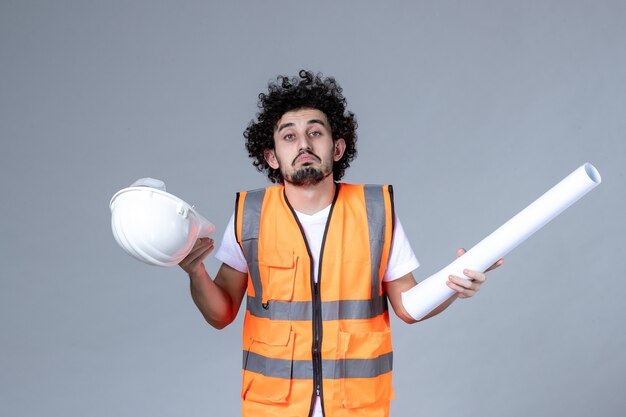 Front view of young unsure uncertain male constructor in warning vest holding safety helmet and showing blank on gray wall