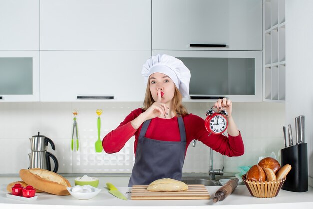 キッチンでハッシュサインを作る赤い目覚まし時計を保持しているエプロンの正面図若い女性