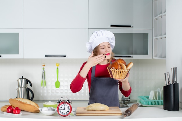 Front view young woman closing eyes in cook hat and apron smelling loaf in the kitchen