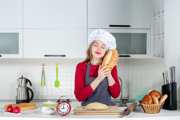 Front view young woman in cook hat and apron closing eyes holding bread in the kitchen