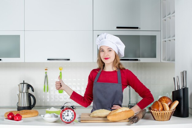 料理の帽子とエプロンでキッチンで親指をあきらめる正面図若い女性