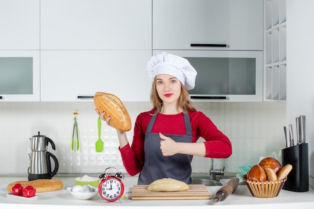 Вид спереди молодая женщина в шляпе повара и фартуке держит хлеб, делая большой палец вверх знак на кухне
