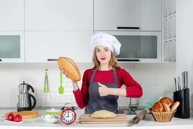 Front view young woman in cook hat and apron holding bread making thumb up sign in the kitchen