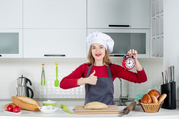 料理の帽子とエプロンでキッチンで親指をあきらめる赤い目覚まし時計を保持している正面図の若い女性