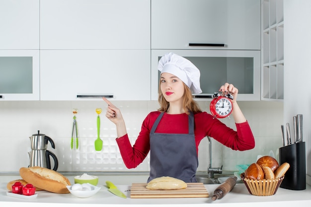 キッチンの左を指す赤い目覚まし時計を保持しているクック帽子とエプロンの正面図若い女性