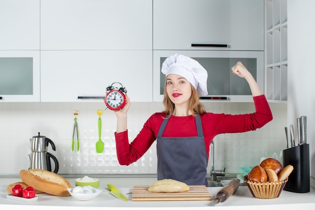 Front view young woman in cook hat and apron holding red alarm clock showing her arm muscle in the kitchen