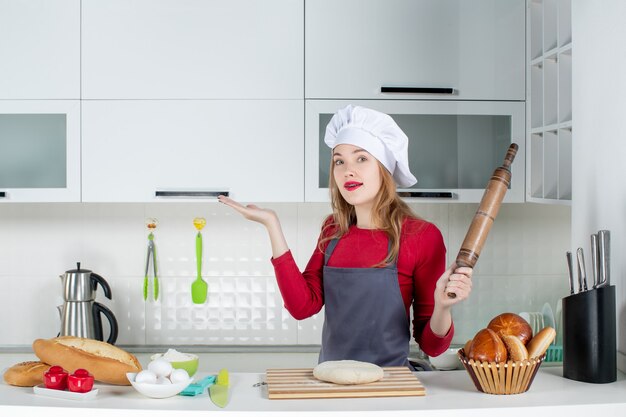 Вид спереди молодая женщина в шляпе повара и фартуке, держащая скалку на кухне