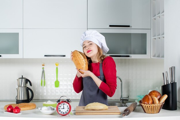 Front view young woman in cook hat and apron holding up bread in the kitchen