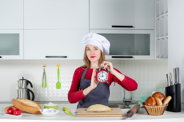Front view young woman in cook hat and apron holding up red alarm clock in the kitchen