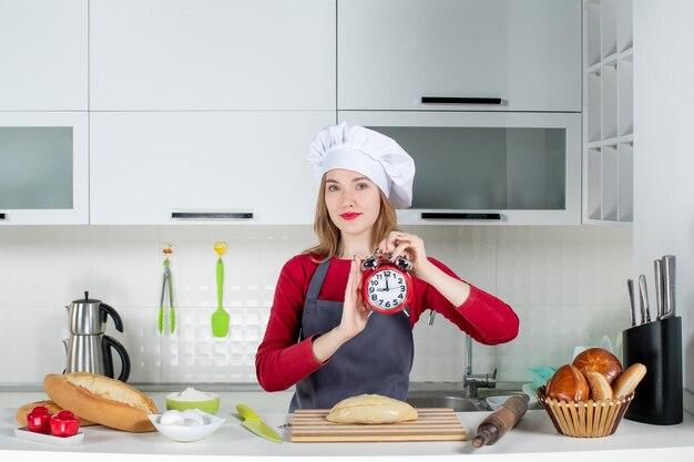 料理の帽子とエプロンでキッチンで赤い目覚まし時計を保持している正面図の若い女性