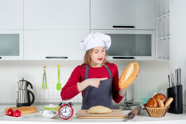 Front view young woman in cook hat and apron pointing at bread in the kitchen