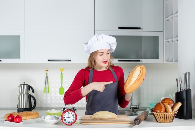 부엌에서 빵을 가리키는 앞치마와 요리사 모자를 쓴 젊은 여성