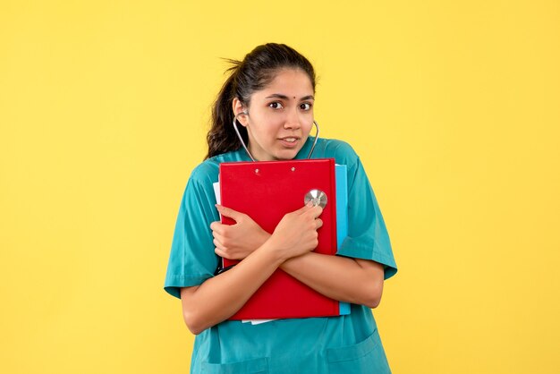 黄色の背景に立っている赤いクリップボードを保持している制服を着た若い女性医師の正面図