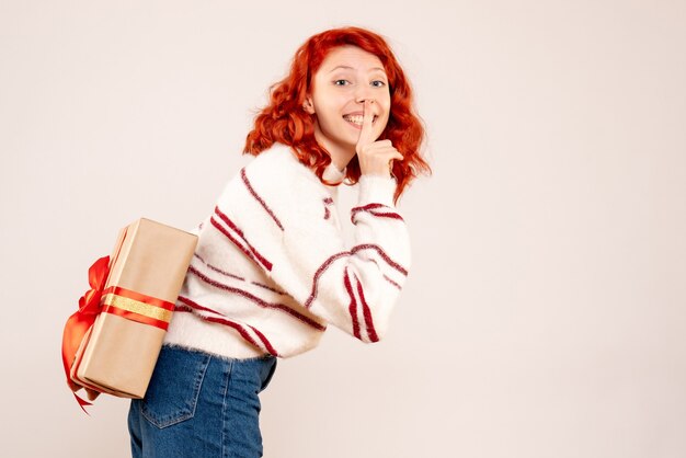 Front view of young woman hiding christmas present behind her back on white wall