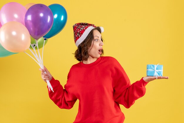 Front view young woman holding balloons and little present on yellow christmas holiday new year emotion color