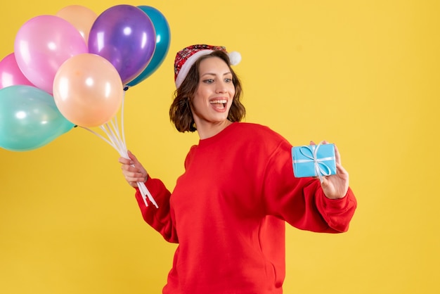 Вид спереди молодая женщина, держащая воздушные шары и маленький подарок на желтом рождественском празднике, новогодняя эмоция женщина