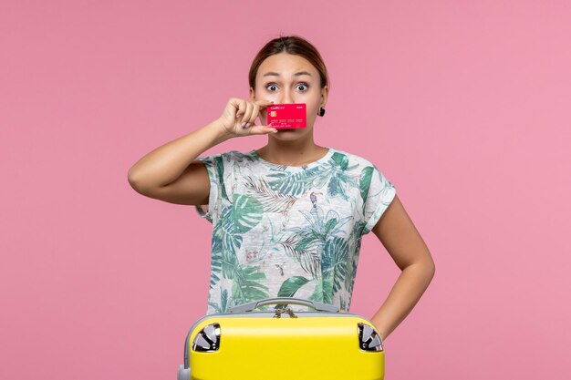 ピンクの壁の飛行航海飛行機の残りの女性に黄色の休暇バッグと銀行カードを保持している正面図若い女性