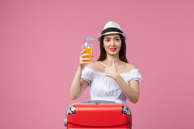 ピンクの壁の休暇の飛行機の航海夏の感情旅行で赤い休暇バッグとカクテルを保持している正面図若い女性