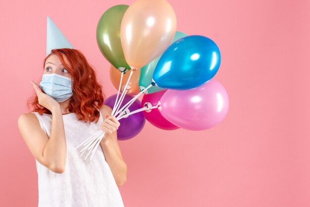 Front view of young woman holding colorful balloons in sterile mask on pink wall