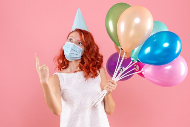 Front view of young woman holding colorful balloons in sterile mask on pink wall