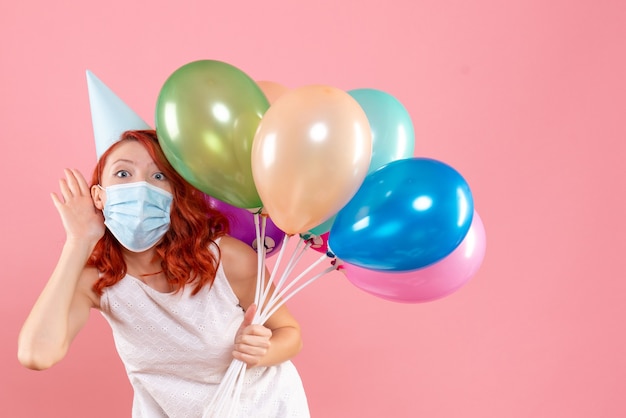 Front view of young woman holding colorful balloons in sterile mask on pink wall