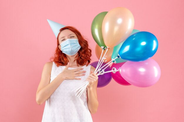 Front view of young woman holding colorful balloons in sterile mask on the pink wall