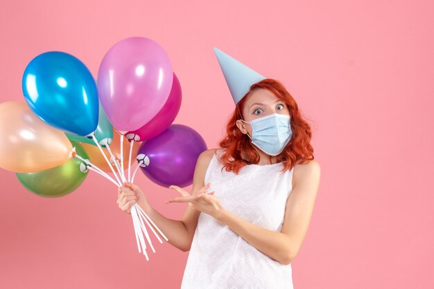 Front view of young woman holding colorful balloons in sterile mask on the pink wall