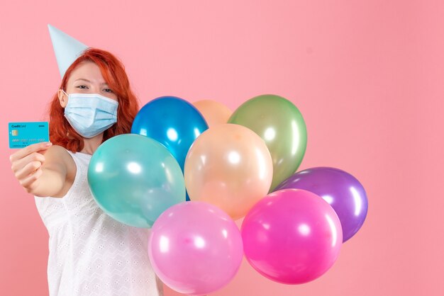 Front view of young woman holding colorful balloons in sterile mask with bank card on pink wall