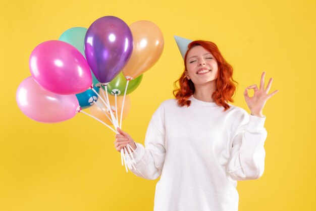 Front view of young woman holding colorful balloons on a yellow wall