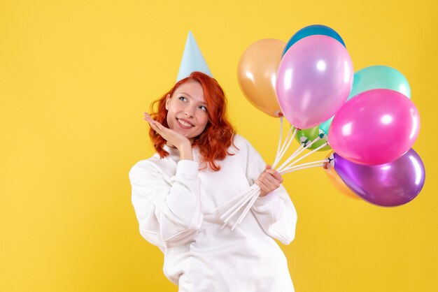 Front view of young woman holding colorful balloons on the yellow wall