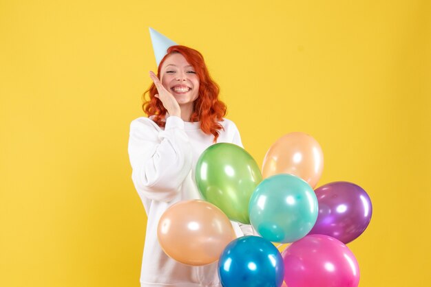 Front view of young woman holding colorful balloons on yellow wall