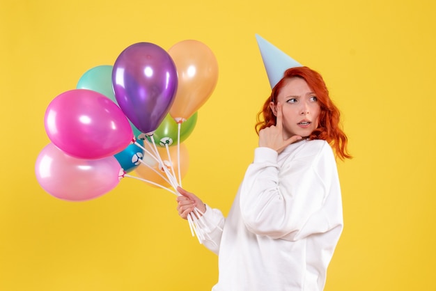 Front view of young woman holding colorful balloons on yellow wall