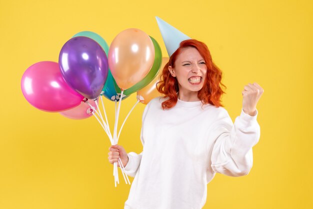 Front view of young woman holding colorful balloons on yellow wall