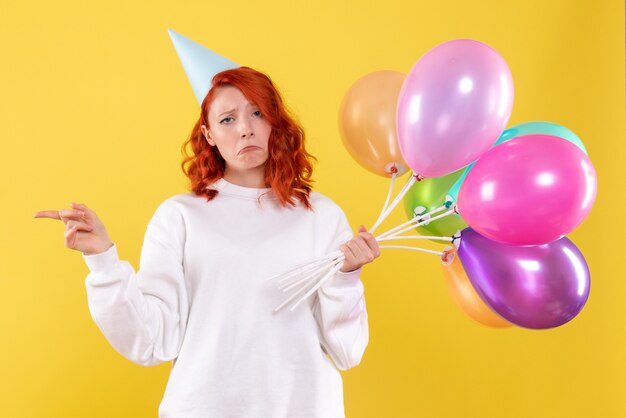 Front view of young woman holding colorful balloons on yellow wall