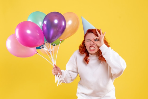 Front view of young woman holding colorful balloons on yellow wall