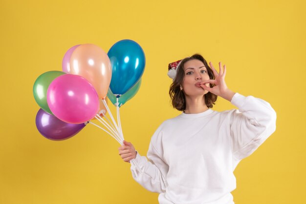 Front view of young woman holding colorful balloons on yellow wall